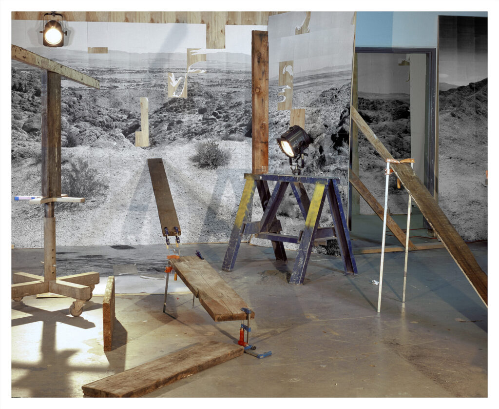 Color image of studio-like space featuring a black and white desert landscape image surrounded by stage lights, wooden boards held in various configurations by clamps.