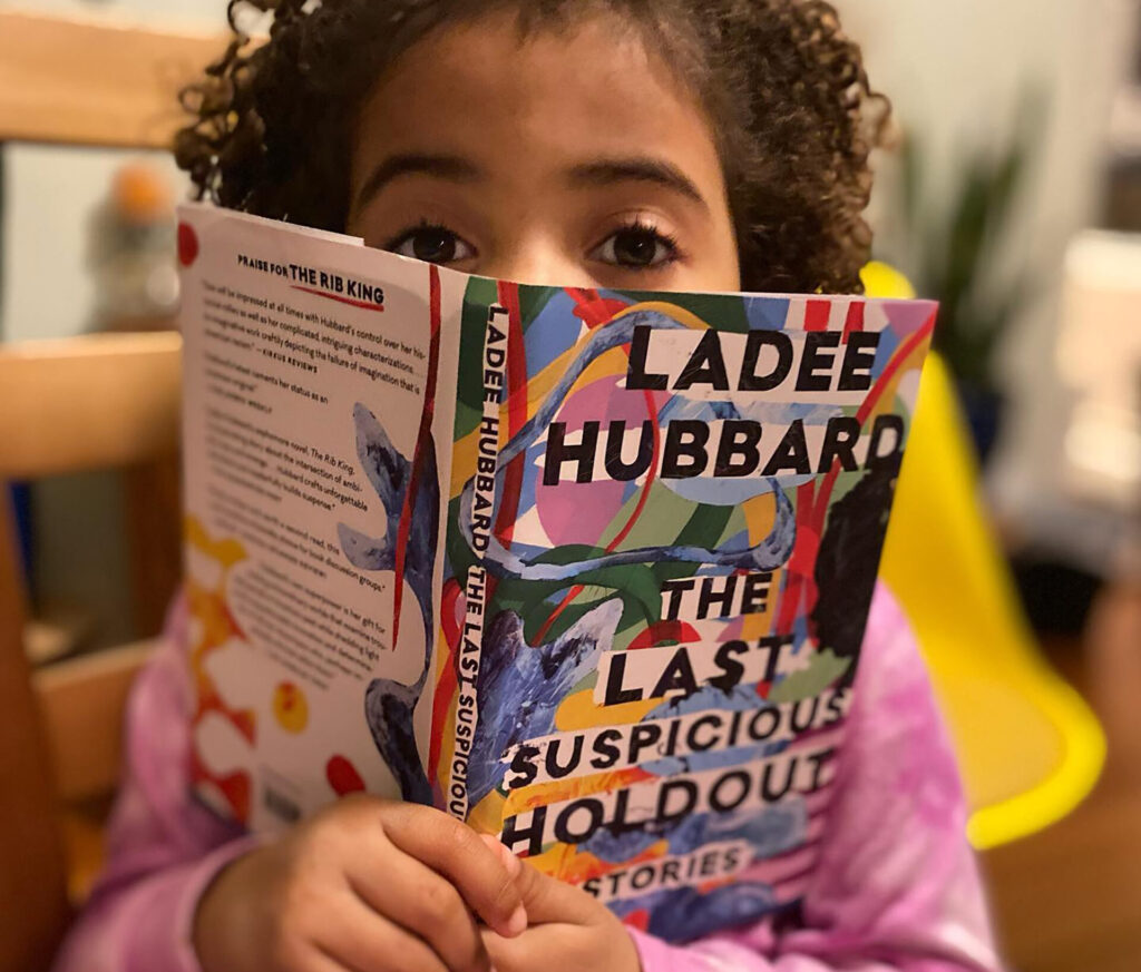 A photo of a child holding a book, The Last Suspicious Holdout by Ladee Hubbard, in front of their face.