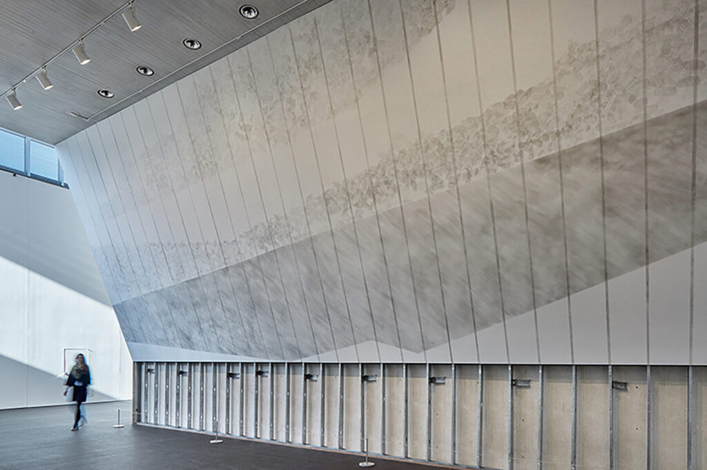 A large tilted wall in a vast hallway; a drawing of lines and round rock shapes covers the wall.