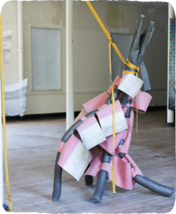 A sculpture of grey tube-like legs draped with pink and white material, leaning, held by a length of yellow strap attached off screen