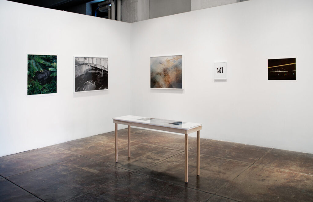 An exhibition view of a table in a room with photos on the walls