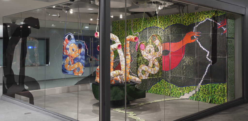 A view of an installation with painted backdrop featuring arms and hands, a painting of tube like forms, and sculptures of tube like forms.
